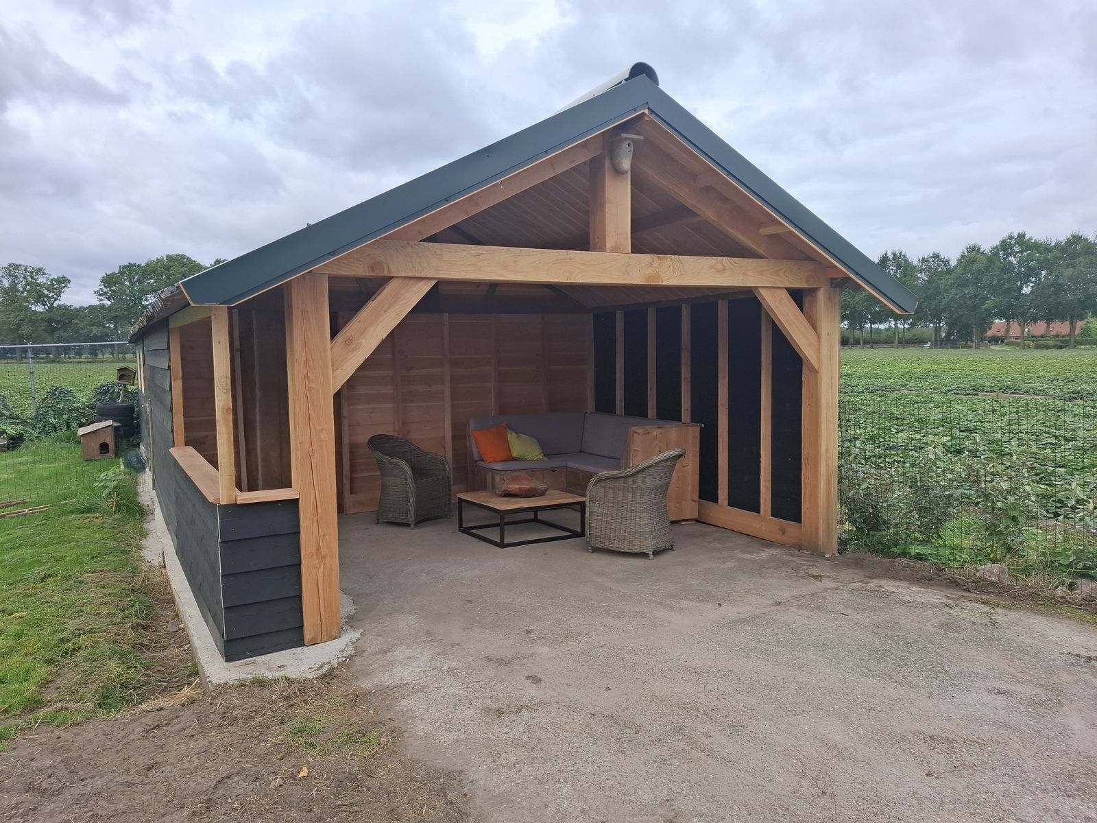 Houten overkapping met loungebank en rieten stoelen, naast de hoeve